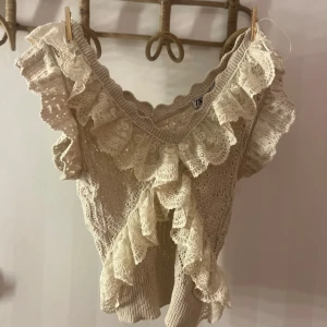 Beige topp med spetsdetaljer från Zara - Säljer en söt beige topp från Zara med vackra spetsdetaljer och volanger. Toppen har en v-ringad design och är ärmlös, perfekt för en romantisk look. Passar bra till både jeans och kjol.