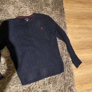 Stickad tröja Ralph Lauren - Säljer en snygg mörkblå stickad tröja från Ralph Lauren med kabelstickat mönster. Tröjan har en liten röd logotyp på bröstet och ribbade kanter vid ärmslut och nederkant. Perfekt för kyligare dagar! Inga defekter alls! Sitter som en en M