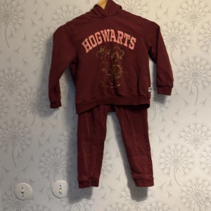 Vinröd hoodie och byxor med Hogwarts-tryck - Säljer ett set med vinröd hoodie och matchande byxor. Hoodien har ett stort Hogwarts-tryck i guld och rosa på framsidan. Perfekt för alla Harry Potter-fans! Setet är bekvämt och passar bra för mysiga dagar hemma.