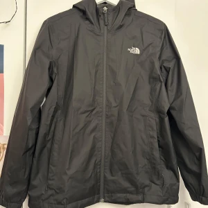 Svart vindjacka från The North Face - Säljer en svart vindjacka från The North Face med huva och dragkedja. Jackan har en stilren design med logotyp på bröstet och är perfekt för blåsiga dagar. Den är lätt och smidig att bära med sig.