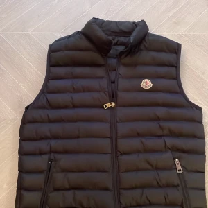 Svart dunväst från Monc - Säljer en stilren svart dunväst från Monc med dragkedja och broderad logotyp på bröstet. Västen har en quiltad design och är perfekt för kyligare dagar. Den är både snygg och praktisk med sina dragkedjefickor.