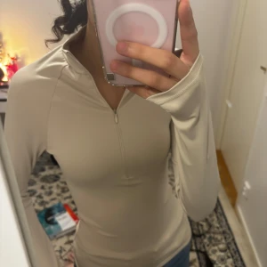 Beige långärmad träningströja Nelly  - Säljer en stilren beige långärmad tröja med dragkedja framtill. Perfekt för en casual look och passar bra till jeans. Tröjan har en tight passform och är tillverkad i ett mjukt material. ALLDRIG använts så gott som ny 