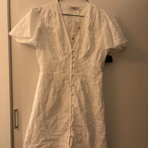 Vit klänning från NA-KD - Säljer en söt vit klänning från NA-KD med korta ärmar och v-ringning. Klänningen har ett broderat mönster och knappar framtill. Perfekt till studenten! 🌸