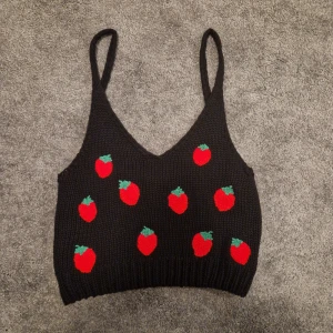 Svart stickad linnetopp med jordgubbsmönster - Säljer en söt svart stickad linnetopp med röda jordgubbar och gröna blad. Toppen har smala axelband och en v-ringad design. Perfekt för en somrig look!