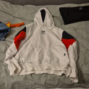 Vit hoodie från Puma - Säljer en vit hoodie från Puma med färgblock i rött och svart på ärmarna. Den har en stor ficka framtill och en bekväm huva. Perfekt för en avslappnad stil.