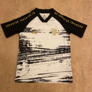Juventus fotbollströja från Adidas 140 - Snygg stilrent Juventus tränings fotbollströja från Adidas med ett svartvitt mönster och guld detaljer. Tröjan har korta ärmar. Perfekt för unga fotbollsfans som äskar Juventus.