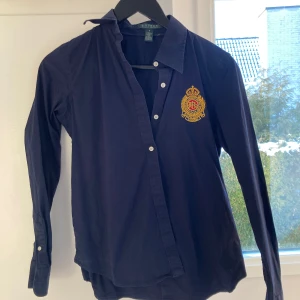 Mörkblå skjorta från Ralph Lauren - Snygg mörkblå skjorta från Ralph Lauren med ett broderat emblem på bröstet. Skjortan är liten storlek 