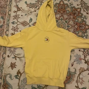 Gul hoodie från Tommy Hilfiger - Säljer en gul hoodie från Tommy Hilfiger med en klassisk logga på bröstet. Den har en känguruficka och en randig detalj på huvan. Perfekt för en avslappnad stil. Ute för ett perfekt pris nu!