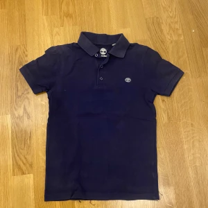 Mörkblå pikétröja från Timberland - Säljer en snygg mörkblå pikétröja från Timberland. Tröjan har en klassisk krage med knappar och korta ärmar. Perfekt för en stilren och avslappnad look. Timberlands logga är broderad på bröstet.