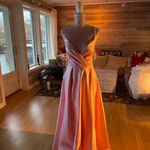 Persikofärgad klänning från JJ's House - Elegant persikofärgad långklänning från JJ's House med omlott-design och smala axelband. Klänningen har en v-ringad urringning och är perfekt för speciella tillfällen. Materialet ger en mjuk och lyxig känsla.