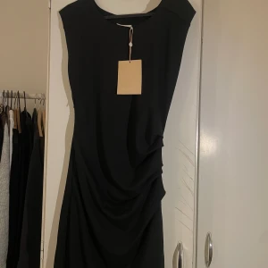Svart klänning från Kaffe - Elegant svart klänning från Kaffe med draperad design på sidan. Klänningen har en ärmlös stil och en klassisk rund halsringning. Perfekt för en stilren look.