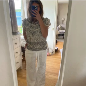 Mönstrad blus med volangdetaljer - Säljer en mönstrad blus med volangdetaljer och korta ärmar. Blusen har ett vackert blommigt mönster i grått och vitt. Perfekt för en stilfull och bekväm look.