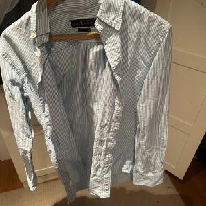 Randig skjorta från Ralph Lauren - Snygg blå- och vitrandig skjorta från Ralph Lauren i klassisk passform. Skjortan har långa ärmar och knappar framtill samt en broderad logotyp på bröstet. Perfekt för en stilren look.