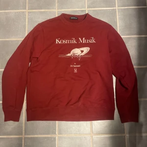 Röd tröja från Undercover - Säljer en röd tröja från Undercover med trycket 'Kosmik Musik' av Jun Takahashi. Tröjan har långa ärmar och en rund halsringning. Perfekt för dig som gillar unika designer och streetwear.