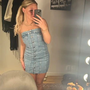 Jeansklänning  - Snygg axelbandslös jeansklänning från gina i ljusblå denim. Klänningen har knappar framtill och en figurnära passform som framhäver silhuetten. Perfekt för en trendig look!