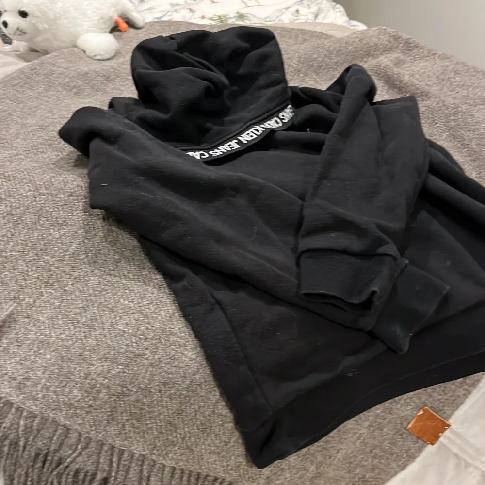 Säljer en stilren svart hoodie från Calvin Klein med logga på bröstet och text på insidan av luvan. Perfekt för en avslappnad look. Hör av dig vid intresse!. Neuletakit & Villapaidat.