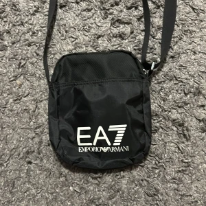Svart  från Emporio Armani - Svart axelväska från Emporio Armani med logotypen EA7 tryckt på framsidan. Väskan har en justerbar axelrem och en praktisk dragkedja för säker förvaring. Perfekt för att bära med sig det viktigaste på ett stilfullt sätt. Bra skick, sällan använd.