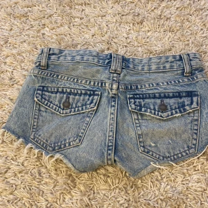 Blå jeansshorts från Pull&Bear - Snygga blå jeansshorts från Pull&Bear med slitna detaljer och fransig kant. Shortsen har en klassisk femficksdesign och knappar framtill. Perfekta för en avslappnad stil.