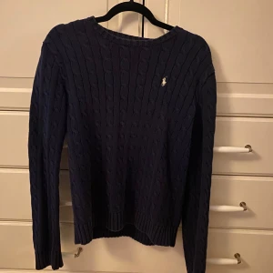 Mörkblå stickad tröja från Ralph Lauren - Säljer en stilren mörkblå stickad tröja från Ralph Lauren med kabelstickat mönster. Tröjan har långa ärmar och en liten logga på bröstet.