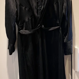 Svart omlottklänning - Elegant svart omlottklänning med knappar framtill och långa ärmar. Klänningen har en stilren design med ett knytband i midjan förgäves en smickrande passform. Perfekt för en chic look.