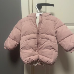 Rosa dunjacka från H&M - Söt rosa dunjacka för små barn från H&M. Jackan har en mysig huva med små öron och är quiltad för extra värme. Perfekt för kyliga dagar med dragkedja framtill för enkel på- och avtagning. Användes bara 2 ggr