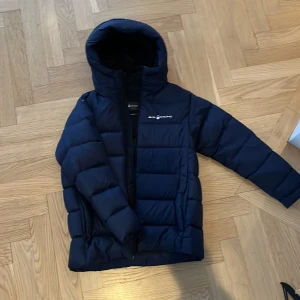 Mörkblå dunjacka från Sail Racing - Säljer en mörkblå dunjacka från Sail Racing. Jackan har en dragkedja framtill och en hög krage med huva. Perfekt för kyliga dagar med sin vadderade design. Skicket är mycket bra på jackan, inga hål eller så men använt ett flertal gånger🫶storleken är 160 men passar på mig som har XS/S.