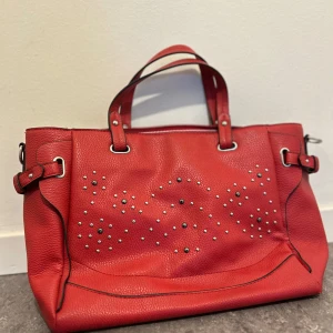 Röd handväska med nitar - Snygg röd handväska i skinn med dekorativa nitar på framsidan. Väskan har två handtag och en rymlig insida med randigt foder och en innerficka med dragkedja. Perfekt för att ge en färgklick till din outfit!❣️