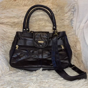 Svart axelväska i skinn - Snygg svart axelväska i skinn med både handtag och justerbar axelrem. Väskan har flera fack och är knappt använd.