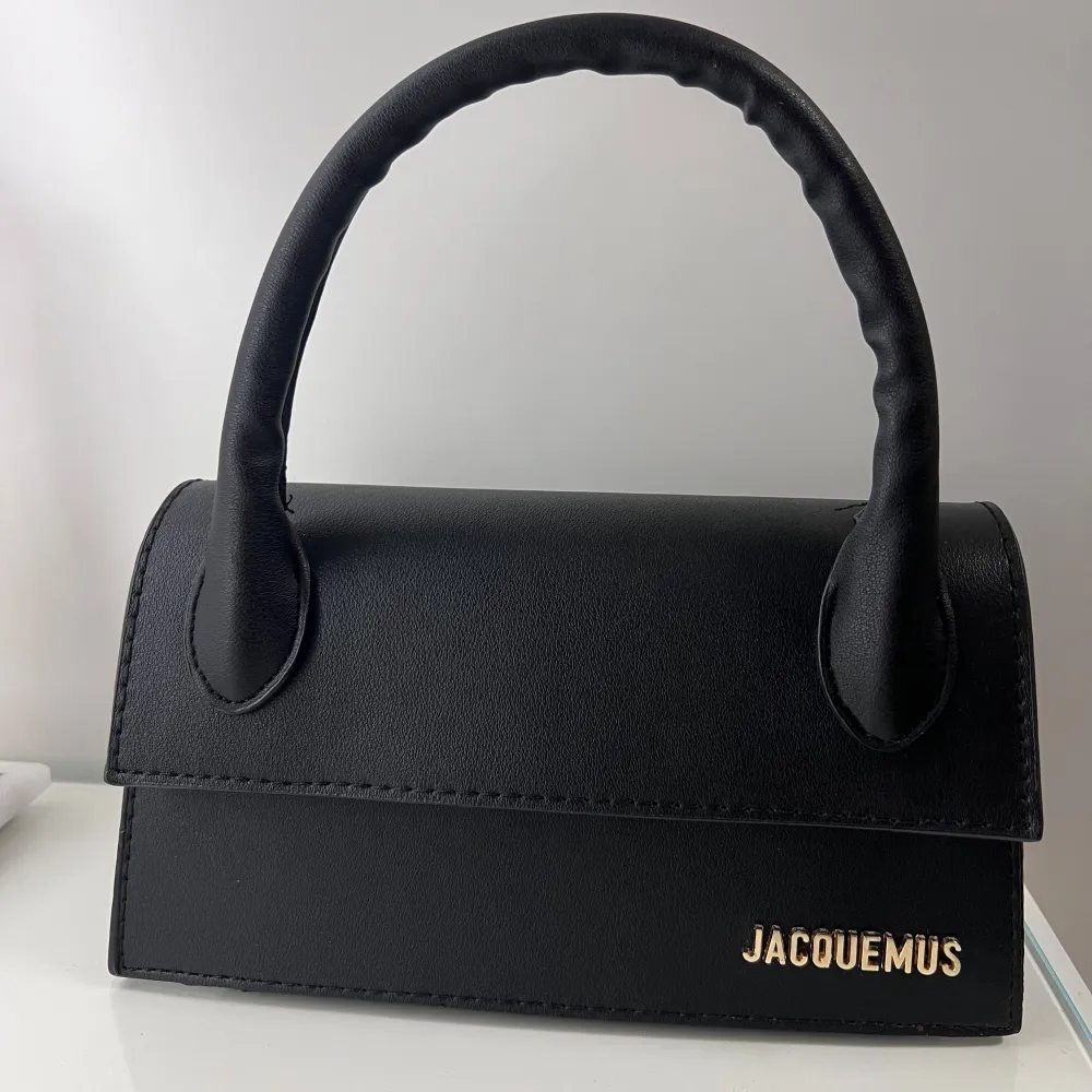 Snygg svart handväska från Jacquemus i syntetmaterial. Väskan har ett stilrent och minimalistiskt utseende med ett handtag och en avtagbar axelrem. Perfekt för att bära dina nödvändigheter med stil. Den har en magnetisk stängning och guldlogotyp på framsidan.. Laukut & Käsilaukut.