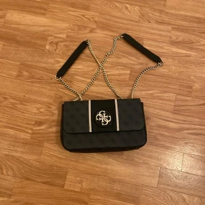 Svart axelväska med kedjedetaljer - Snygg svart axelväska från Guess med guldfärgade kedjor och logotyp på framsidan. Väskan har ett diskret mönster och en praktisk storlek för vardagsbruk. Insidan är fodrad och har flera fack för enkel organisering.