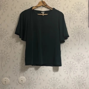 Mörkgrön blus med korta ärmar - Elegant mörkgrön blus med korta, fladdriga ärmar. Perfekt för en stilren look. Blusen har en rund hals och är tillverkad i ett mjukt material.