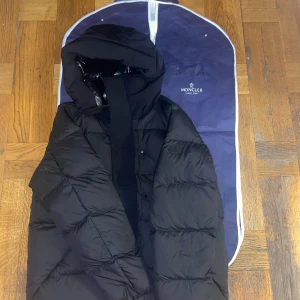 Moncler madiera jacka - Säljer en stilren svart dunjacka från Moncler med huva och dragkedja. Jackan har en quiltad design och är perfekt för kyliga dagar. Moncler-logotypen syns på bröstet och insidan har en glansig finish. Pris kan diskuteras vid snabb affär!!
