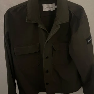 Olivgrön overshirt från Calvin Klein - Snygg olivgrön overshirt från Calvin Klein med knappar framtill och två bröstfickor. Perfekt för lager-på-lager-stil. Klassisk krage och långärmad design gör den mångsidig och stilren. Knappt använd och passar perfekt nu till våren!Mvh Tage