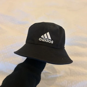 Svart bucket hat från Adidas - Snygg svart bucket hat från Adidas med broderad logga framtill. Perfekt för en avslappnad stil och skydd mot solen. Passar till både vardag och fritid.