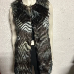 Brun och svart pälsväst - Snygg och trendig pälsväst i bruna och svarta nyanser. Perfekt för att lägga till en lyxig touch till din outfit. Västen är ärmlös och har en mjuk och fluffig textur. Konstpäls från Desires.Storlek S. Helt ny. Ordpris 699kr.