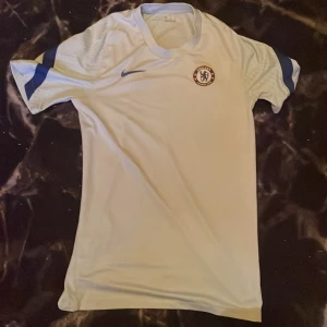 Ljusblå Chelsea fotbollströja från Nike - Säljer en vit Chelsea fotbollströja från Nike med korta ärmar och blå detaljer. Tröjan har Dri-FIT-teknologi för optimal komfort och klubbens emblem på bröstet. Perfekt för alla Chelsea-fans! ⚽