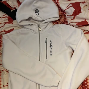 Vit hoodie från Sail Racing - Snygg vit hoodie från Sail Racing med dragkedja och logotyp på bröstet. Den har en justerbar huva med tryck och en praktisk bröstficka med dragkedja. Perfekt för en stilren och sportig look. Storlek 150 men passar även XS/S (köpte för 1000kr)