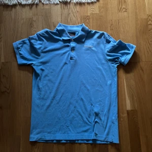 Blå pikétröja från Kappa - Säljer en snygg blå pikétröja från Kappa. Tröjan har en klassisk krage med två knappar och Kappa-loggan broderad på bröstet. Perfekt för en avslappnad stil.