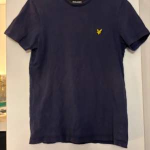 Mörkblå t-shirt från Lyle & Scott - Snygg mörkblå t-shirt från Lyle & Scott med korta ärmar och ett broderat gult emblem på bröstet. Perfekt för en avslappnad stil.