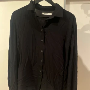 Svart blus från NA-KD - Elegant svart blus från NA-KD med knappar framtill och långa ärmar. Blusen har en stilren design med knytband vid ärmsluten för en extra touch. Perfekt för en chic look. Oversized. 