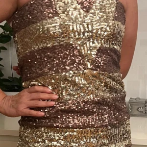 Glittrig paljettklänning - Snygg och glittrig klänning med paljetter i guld och brunt. Den har en tight passform och är ärmlös, perfekt för festliga tillfällen. Klänningen är kort och har ett iögonfallande mönster som verkligen sticker ut.