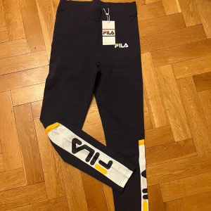 Svarta leggings från FILA - Snygga svarta leggings från FILA med vita och gula detaljer längs benen. Perfekta för träning eller en avslappnad dag. De har en hög midja och är tillverkade i ett stretchigt material för bästa komfort.