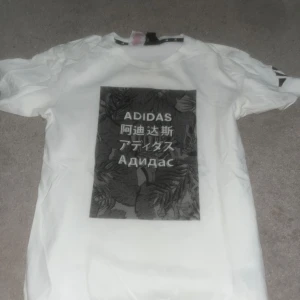 Vit t-shirt från Adidas med tryck - Snygg vit t-shirt från Adidas med ett stort svart tryck på framsidan som har text på flera språk. Perfekt för en sportig look. Kortärmad och tillverkad i bomull för en bekväm passform. 