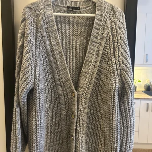 Grå stickad kofta från KappAhl - Mysig grå stickad kofta från KappAhl med knappar framtill. Perfekt för kyliga dagar med sin långa och bekväma passform. Ett måste i garderoben för en stilren look.