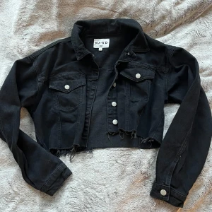 Svart croppad jeansjacka från NA-KD - Snygg svart croppad jeansjacka från NA-KD med knappar framtill och två bröstfickor. Jackan har en rå kant nedtill och långa ärmar med knappdetaljer vid ärmsluten. Perfekt för en utekväll på sommaren eller bara ha i vardagen!💋
