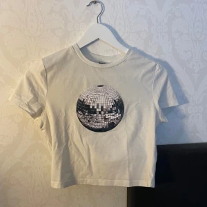 Vit croppad t-shirt med discokula - Snygg vit croppad t-shirt med ett stort tryck av en discokula på framsidan. Perfekt för en avslappnad stil med en touch av festkänsla. Kortärmad och i mjukt material.