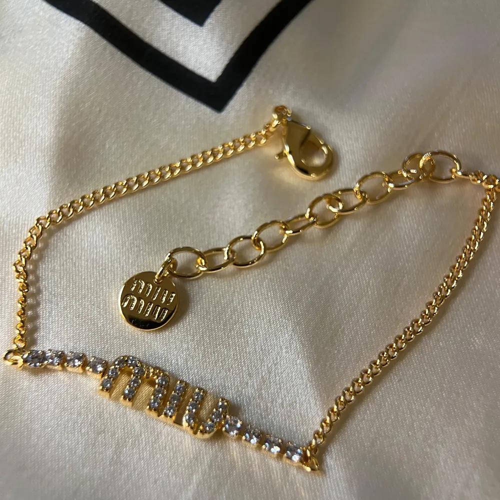 Elegant guldarmband från Miu Miu med glittrande strassdetaljer. Armbandet har en justerbar kedja och en liten rund berlock med ingraverad text. Perfekt för att ge en lyxig touch till din stil.. Asusteet.