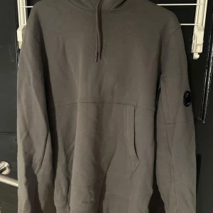 Grå hoodie från C.P. Company - Säljer en stilren grå hoodie från C.P. Company. Tröjan har en klassisk design med en justerbar huva och en ficka framtill. Märkets logotyp finns på ärmen som en snygg detalj.
