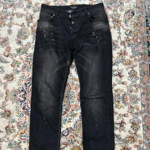 Svarta jeans från Philipp Plein - Snygga svarta jeans från Philipp Plein med coola detaljer som små vita stänk och broderad logga på bakfickan. Byxorna har en klassisk femficksdesign och knappar med märkets namn. Perfekta för en stilren look.