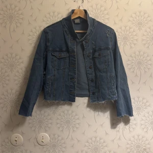 Blå jeansjacka - Säljer en cool blå jeansjacka med slitna detaljer och knappar framtill. Jackan har en croppad design och långa ärmar. Perfekt för en avslappnad stil.